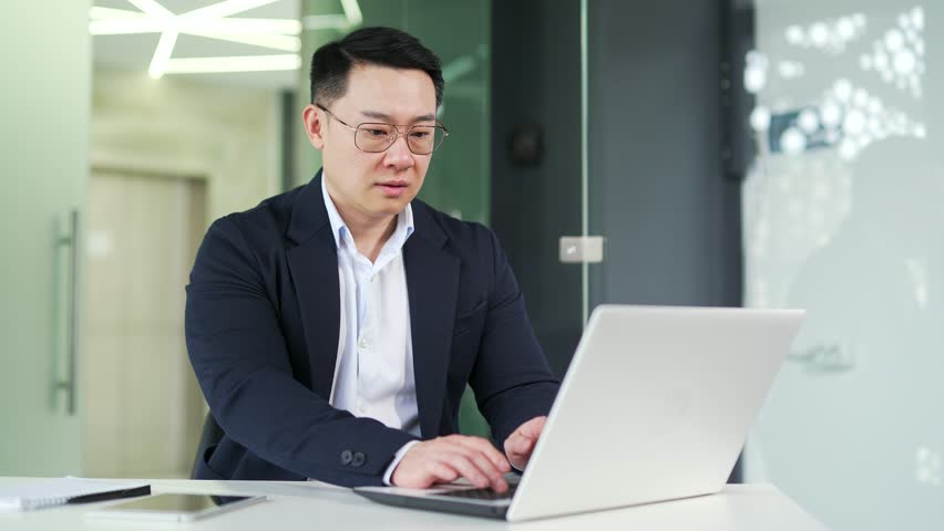 Asian man suffering from back pain while working on laptop while sitting at desk at workplace in modern office Sick employee in a formal suit has back muscle spasms, massages his lower back, stretches Royalty-Free Stock Footage #1103553895