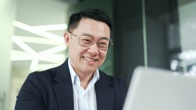 Close up of smiling asian businessman in formal suit working typing on laptop while sitting at desk at workplace in modern office. Happy entrepreneur looks at or writes a message to a client by email - Powered by Shutterstock - Get 15% off with code: PIKWIZARD15