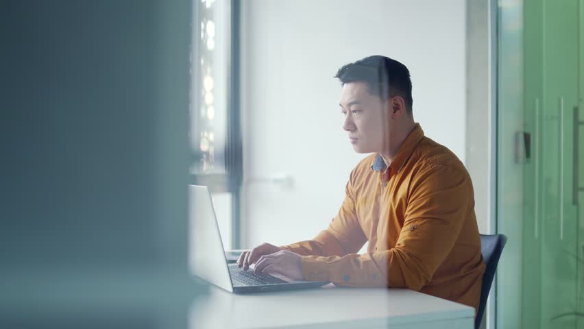 Side view Thoughtful asian businessman working typing on laptop while sitting at desk at workplace in modern office. Silhouette serious employee browses pc messages, chats online with a client Royalty-Free Stock Footage #1103553921