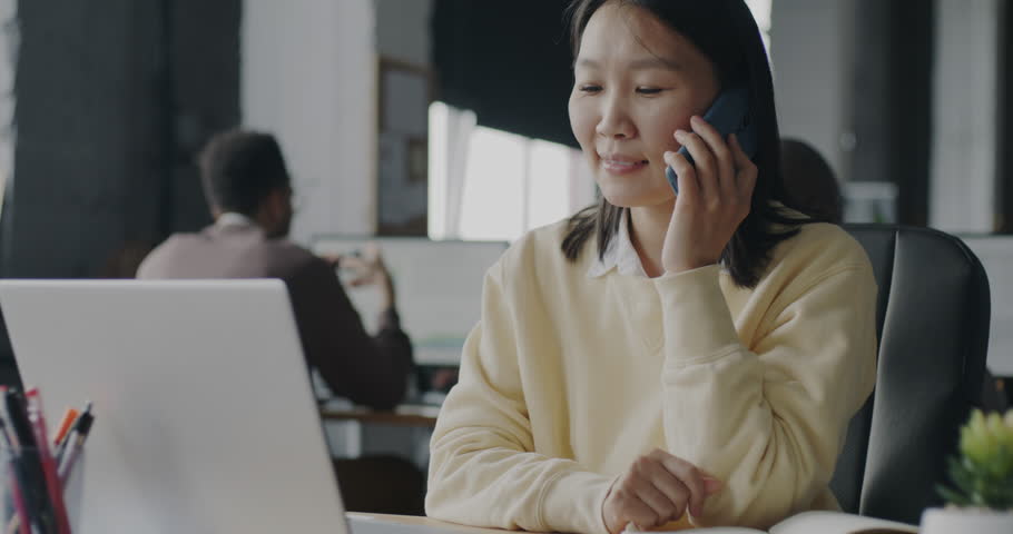 Young Asian lady office worker speaking on mobile phone and typing with laptop working indoors. Colleagues involved in business activities in background. Royalty-Free Stock Footage #1103555595