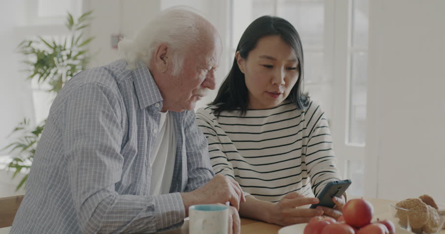 Young woman teaching senior father how to use smartphone in apartment in kitchen. People talking and touching device screen discussing modern technology.
