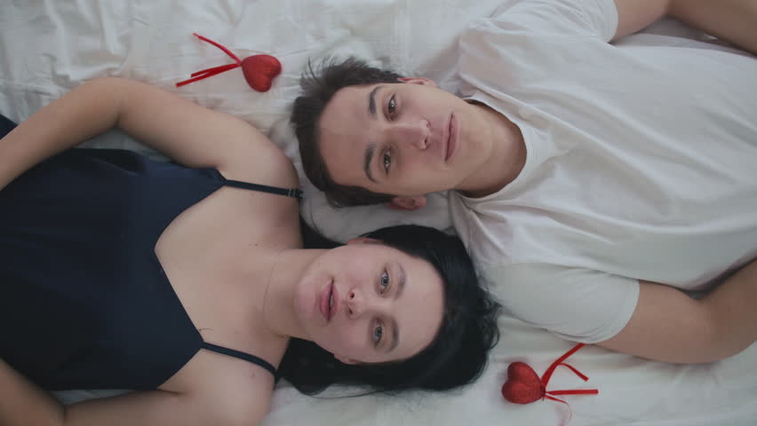 Top view young couple with serious facial expression lying on bed looking at camera and looking at each other. Portrait of Caucasian handsome man and beautiful woman posing on Valentine