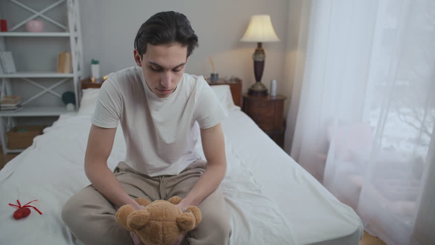 Portrait of depressed lonely Caucasian young man with teddy bear sitting on bed thinking. Front view abandoned frustrated guy with toy alone at home indoors | Shutterstock HD Video #1103555943