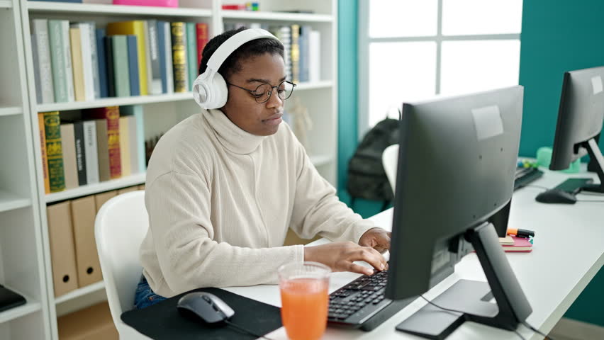 African american woman student using computer and headphones drinking orange juice at library university
