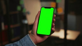 Footage of modern smartphone in male hands isolated on blurred background of office in evening. Close-up shot of unrecognizable man using gadget with green screen. Chroma key concept. Indoors