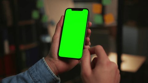 Footage of modern smartphone in male hands isolated on blurred background of office in evening. Close-up shot of unrecognizable man using gadget with green screen. Chroma key concept. Indoors : vidéo de stock