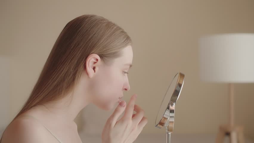 Worried young woman looks in the mirror, examining her facial skin. Unhappy woman Generation Z, dissatisfied with her skin condition, thinks about professional skin care products. Skin care concept ad | Shutterstock HD Video #1103564733