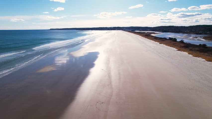 Beach flyover into a bright sunny sky with tides on the sand and waves crashing. Ogunquit beach in south Maine, New England beachside on Atlantic ocean.