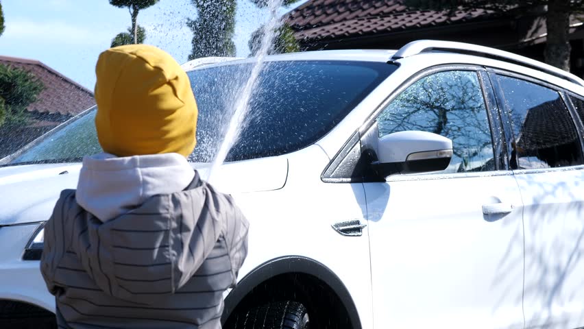 a boy pours water from a garden hose onto a car, a boy washes a white car