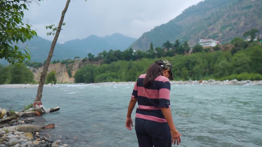 Rear view of Indian teenager girl traveler wearing cap and open arms while standing near Beas river at Manali, Himachal Pradesh, India. Female in background of mountain and river 