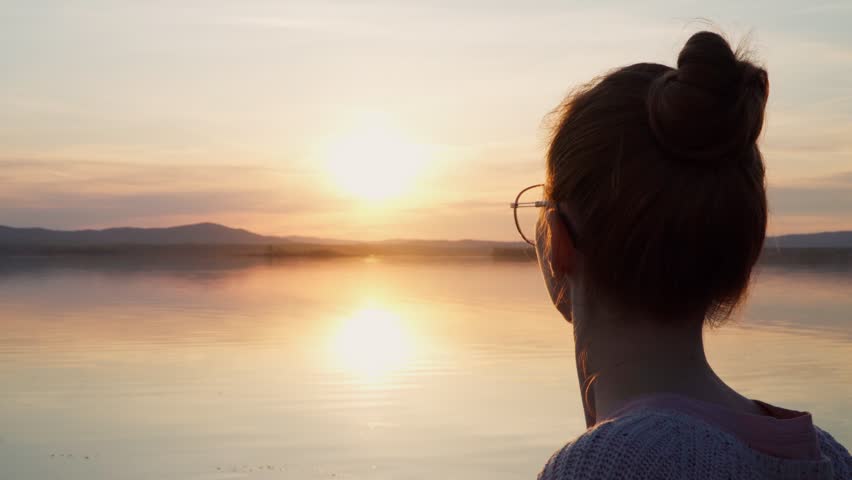 A girl with glasses looks at the sunset on the shore of a calm lake. Mosquitoes fly around trying to bite. Royalty-Free Stock Footage #1103575951