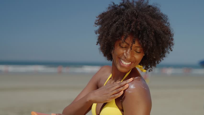 Close up of a happy smiling young black woman is applying a sunscreen or sun tanning lotion on a shoulder to take care of her skin on a seaside beach during holidays vacation.
 Royalty-Free Stock Footage #1103576033