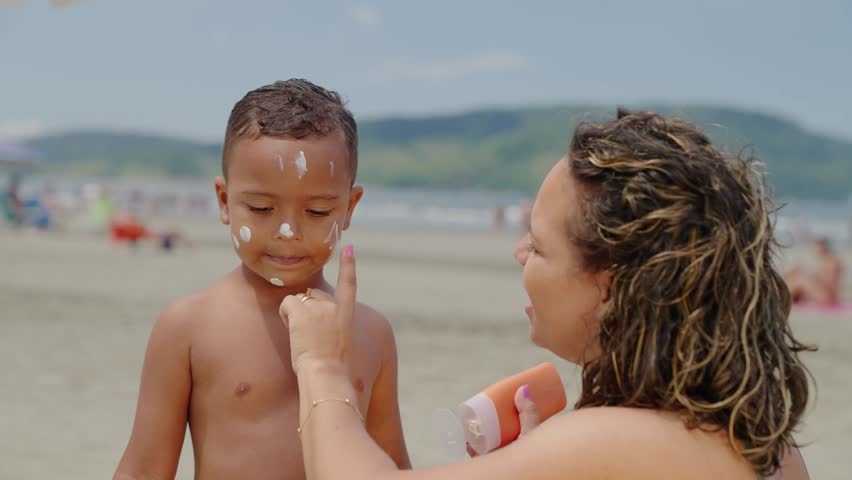 mother applying sunblock lotion on sons skin for sun protection little boy getting ready to swim on the beach with mom using sunscreen caring for childs health on sunny day 
 Royalty-Free Stock Footage #1103576041