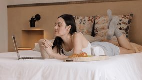 Caucasian positive young woman lying on bed, making online video call with laptop, speaking and gesturing at home in the morning. Indoors. High quality 4k footage