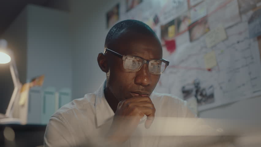 Tired black detective reading report, taking off eyeglasses and rubbing his nose bridge, working late on crime investigation in his office during night. Medium shot, focus on foreground Royalty-Free Stock Footage #1103589981