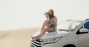 A young beautiful woman of European appearance shoots a video sitting on the hood of a car among the sands in the desert