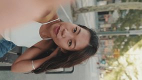 Vertical video, Pretty brunette girl, dressed in white top and jeans, smiles making video call from smartphone while sitting on bench on cityscape background, waving her hand in greeting