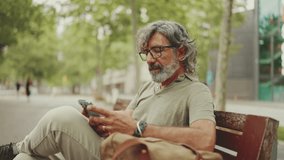 Positive middle-aged man with gray hair and beard wearing casual clothes sits on bench and uses mobile phone. Mature gentleman in eyeglasses writes message on smartphone - Powered by Shutterstock - Get 15% off with code: PIKWIZARD15