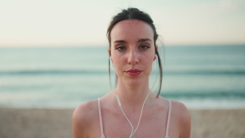Young athletic woman with long ponytail wearing beige sports top in wired headphones, smiling at the camera while standing on the sea background Royalty-Free Stock Footage #1103603125