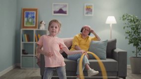 Pretty granddaughter turns hula hoop while nanny listen to audio-book on sofa at home. Hyperactive child under careful granny control. Family lifestyle - Powered by Shutterstock - Get 15% off with code: PIKWIZARD15
