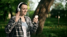 Young woman in casual checkered shirt and jeans in city summer park outdoors, talk by video call mobile cell phone spread hand