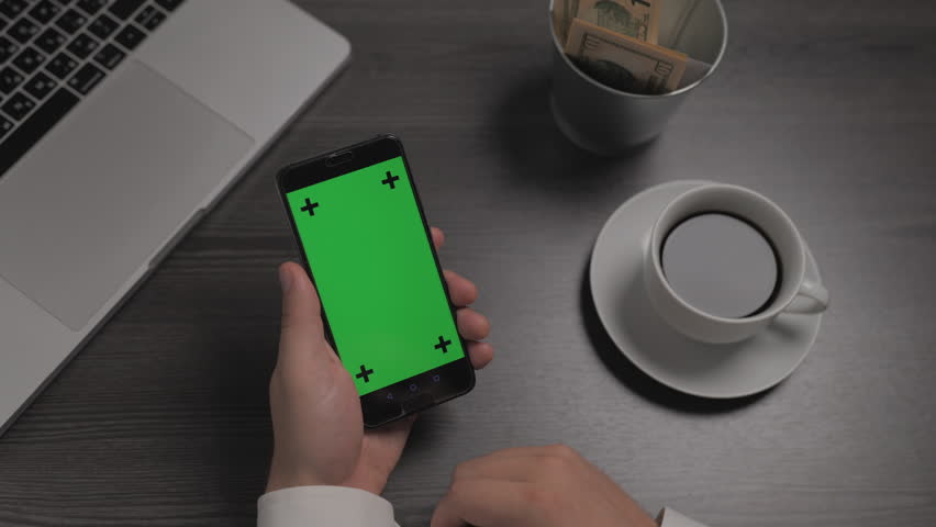 Mobile phone with green screen chroma key laying near on the table. Businessman hands drinking coffee, swipe and tousch a mobile phone screen. Digital technology, business, cyberspace device concept