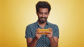 Worried funny addicted indian man enthusiastically playing racing video games on mobile phone. Handsome young guy using smartphone gadget app with drive simulator isolated on yellow studio background