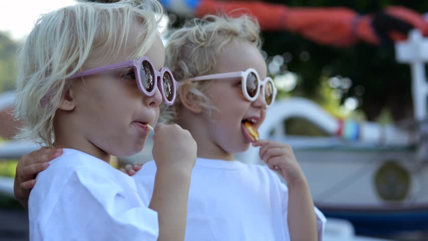 Close-up slow-motion shot of two girls kids eating lollipops in nature in hot sunny weather and enjoying view. Two twin sisters eat lollipops in nature and point their fingers at interesting views