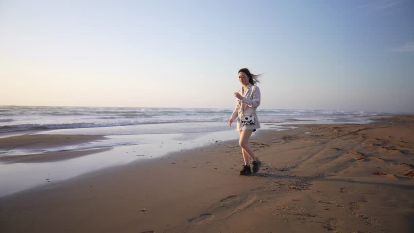 Young woman in white jacket and black shorts walking on beach at sunset 