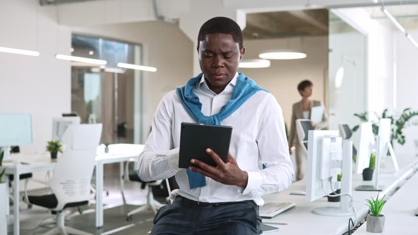 Handsome African American trader sitting on table in office. Hardworking man using tablet computer tapping on sensor screen. In background walking workers with technology devices. Work process.