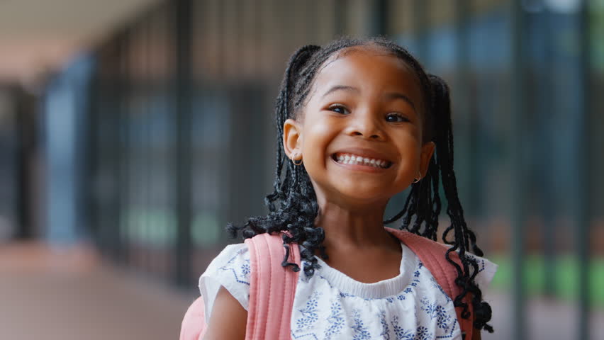 Head and shoulders portrait of smiling female elementary school student wearing backpack standing outside school building - shot in slow motion