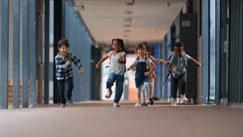 Handheld shot showing class of multi-cultural elementary school students running along outside walkway by school building - shot in slow motion 库存视频
