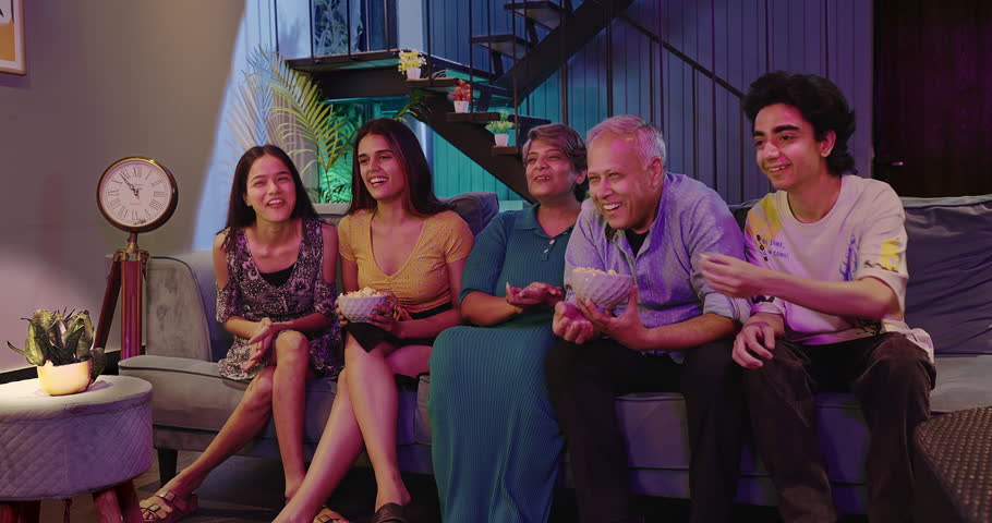 Happy Indian family watching tv with grandparents eating snack young teen girl laugh hold popcorn bowl enjoy television film at home. Group of people sitting on sofa having fun joy spend time together
