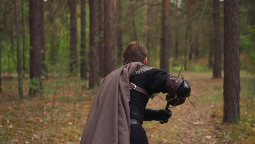Strong knight with cape flying in wind holds sword running through forest backside view slow motion. Man takes part in medieval role-playing reconstruction