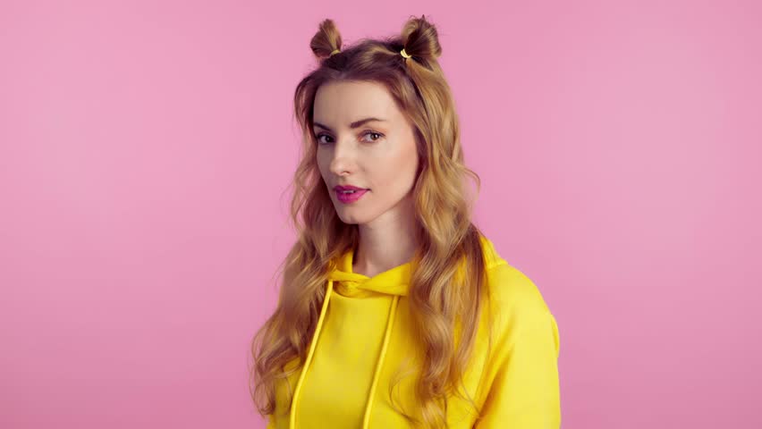 Woman sends air kiss smiling looking at camera. Happy positive emotional blonde girl. Joyful cheerful pretty lady in yellow hoodie, smile face. Indoors studio, isolated on pink background | Shutterstock HD Video #1103656597