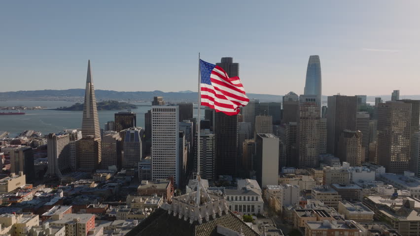 Slide and pan footage of US flag waving in wind on top of building above city. Modern downtown skyscrapers in background. San Francisco, California, USA Royalty-Free Stock Footage #1103657261
