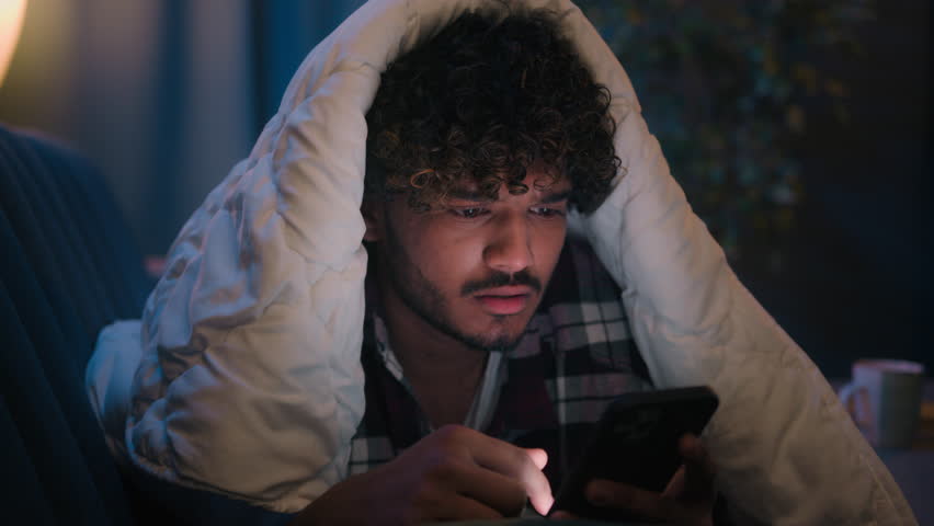 Shocked guy Indian man Arabian male shock wonder reading news in phone social media amazed with smartphone message at night evening home on couch under blanket cover with duvet surprised gadget addict Royalty-Free Stock Footage #1103658769