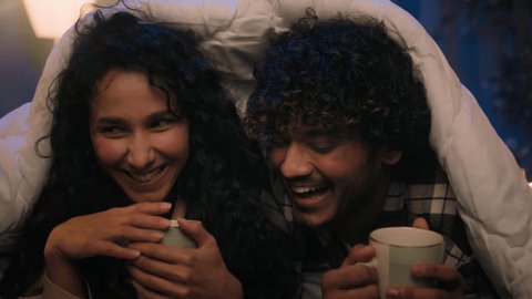 Multiracial couple love diverse boyfriend and girlfriend man and woman drink tea cups talking laughing cuddling affectionate romantic bonding under warm blanket cover duvet at cozy evening night home Video stock