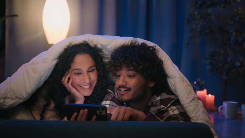 Multiracial couple man and woman at night home using mobile phone shocked amazed with online news shock wonder smartphone social media at evening on couch under blanket cover with duvet gadget addict Royalty-Free Stock Footage #1103658793