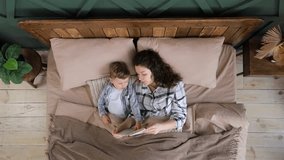 Top view of young woman reads a book with fairy tales to her toddler son before going to bed. Child and his mom in cute pajamas reads a book lying in bed. Happy childhood. Slow motion video