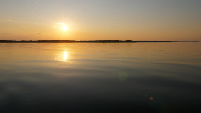 Smooth water of big lake at calm weather, magical sunset hour. Bright sun shine low over horizon, dark stripe of land seen at distance. Quiet and beautiful evening time at Ladoga lake.