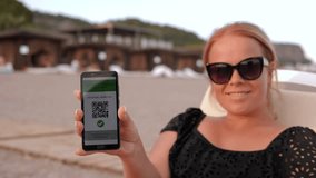 Portrait of a young woman on the beach in a swimsuit and sunglasses, she is holding a phone with a QR code about vaccination against coronavirus infection. - Powered by Shutterstock - Get 15% off with code: PIKWIZARD15
