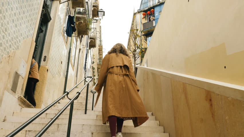 Woman in a raincoat walks up the stairs along a narrow street in Lisbon, Portugal, rear view. The walls are decorated with azulejo tiles. Royalty-Free Stock Footage #1103667771
