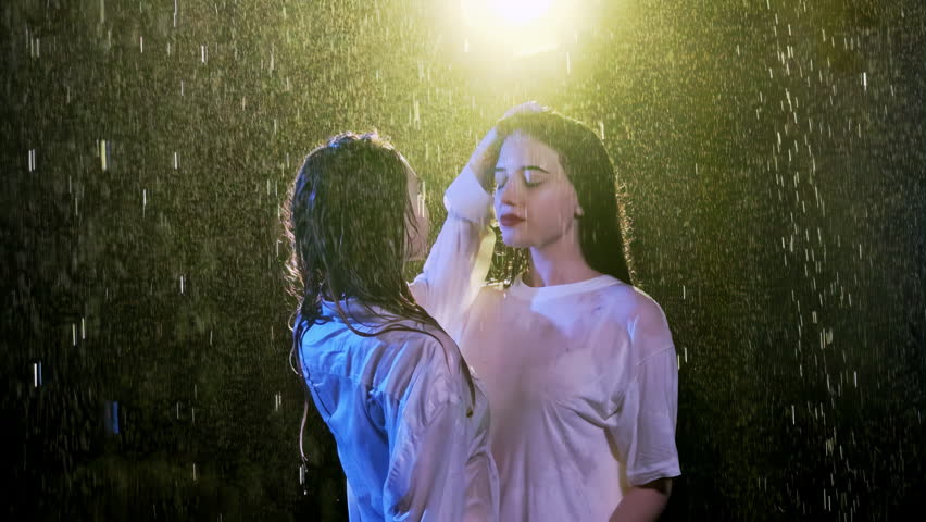 Young woman keeps hand on wet head of brunette with red lipstick on lips. Attractive females stand under pouring rain against lantern