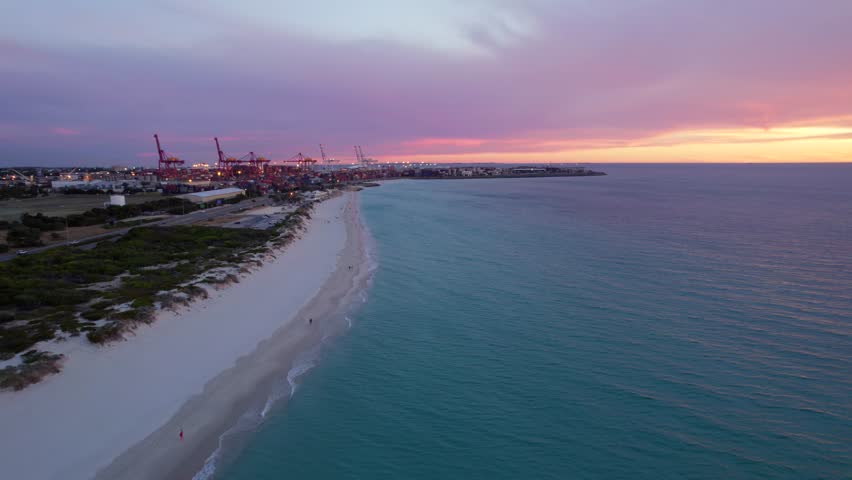 An aerial shot of Port Beach at sunset in Perth, Western Australia. People walk across the beach and watch the sunset. Container shots visable out too sea.