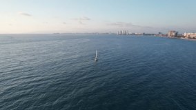 Drone video orbiting around a sailboat cruising along the coast of Puerto Vallarta, showcasing not only the sailboat but also the coast, mountains, and part of the city.