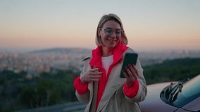 Caucasian smiling hipster girl saying Goodbye at smartphone camera during video call. Attractive millennial female waving and posing to mobile phone while standing near car during travel at sunset