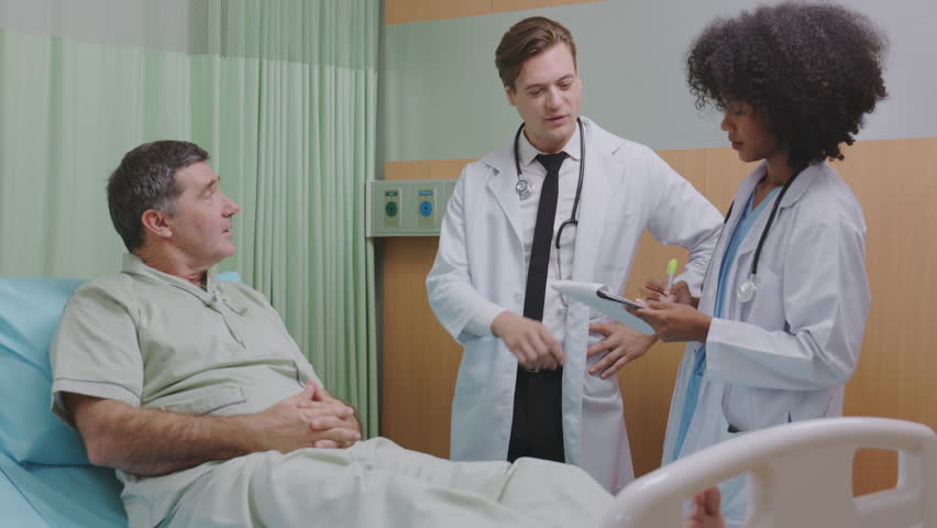 Caucasian male doctor talks to elderly patient lying in bed, while an African female nurse stands taking notes. The friendly conversation reflects their caring approach to the patient's healthcare. Royalty-Free Stock Footage #1103679687