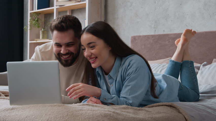 Happy Caucasian woman and man lying on bed carefree couple watch funny video on laptop in bedroom smiling husband boyfriend and wife girlfriend homeowners surfing computer website having fun joy laugh - Powered by Shutterstock - Get 15% off with code: PIKWIZARD15