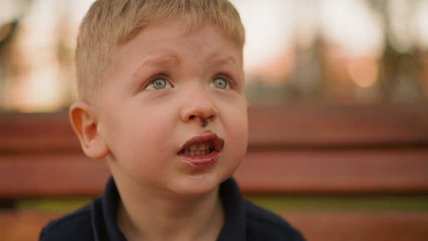 Funny toddler boy licks out chocolate from upper lip in city park at sunset closeup slow motion. Green eyed child with messy face. Sweet tooth in garden
